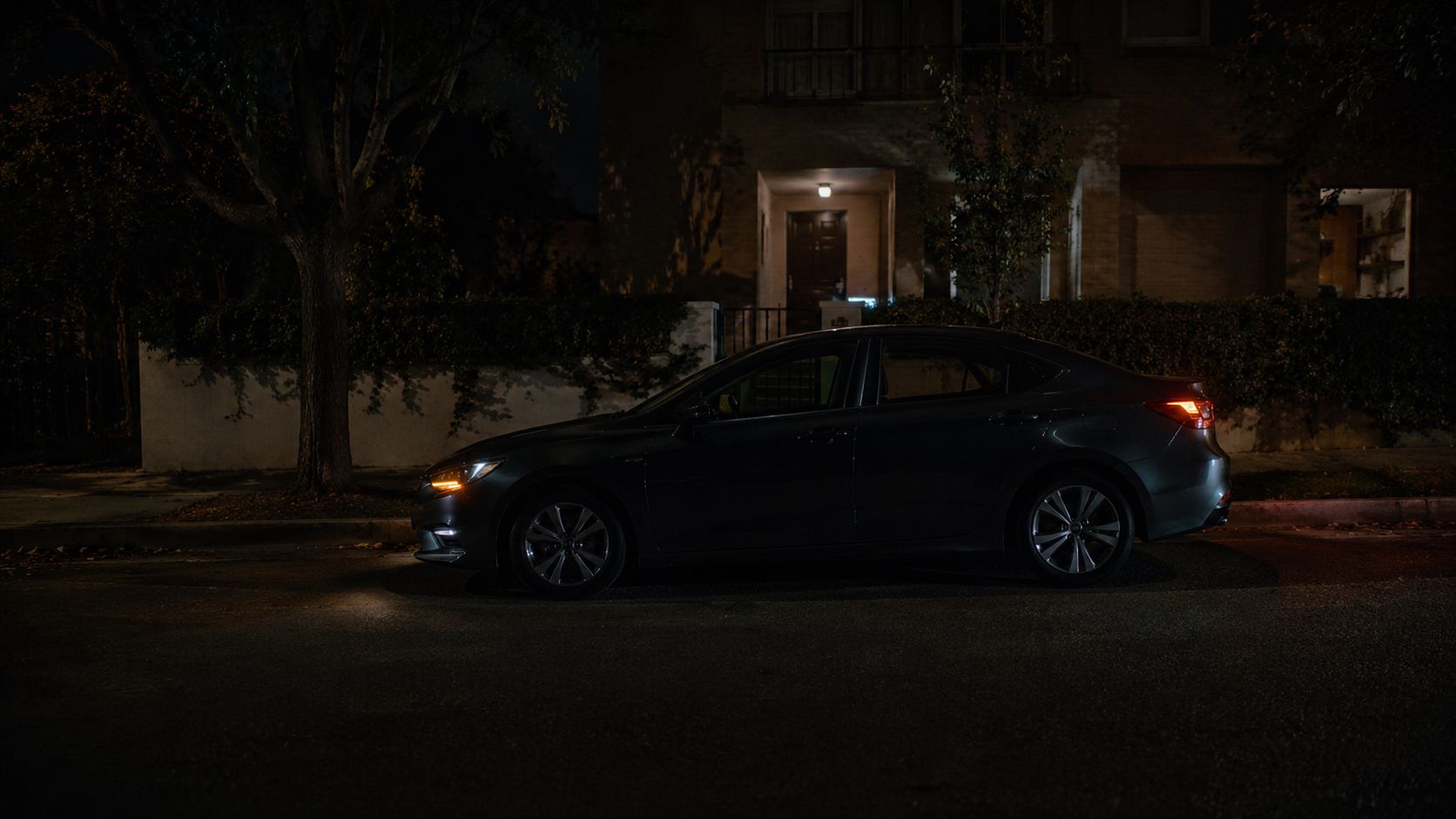 En mørk Mazda sedan parkeret på en vej om natten med tændte positionslys og baglygter i siden.
