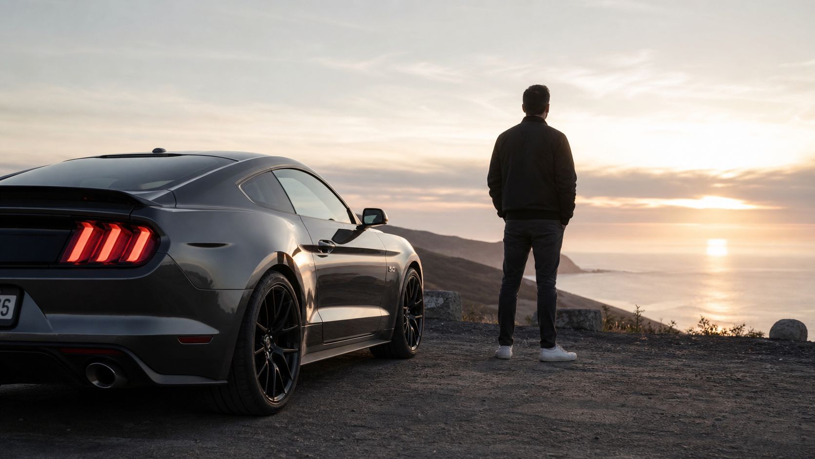 En mand står ved siden af sin mørkegrå Ford Mustang og kigger ud over havet ved solnedgang.