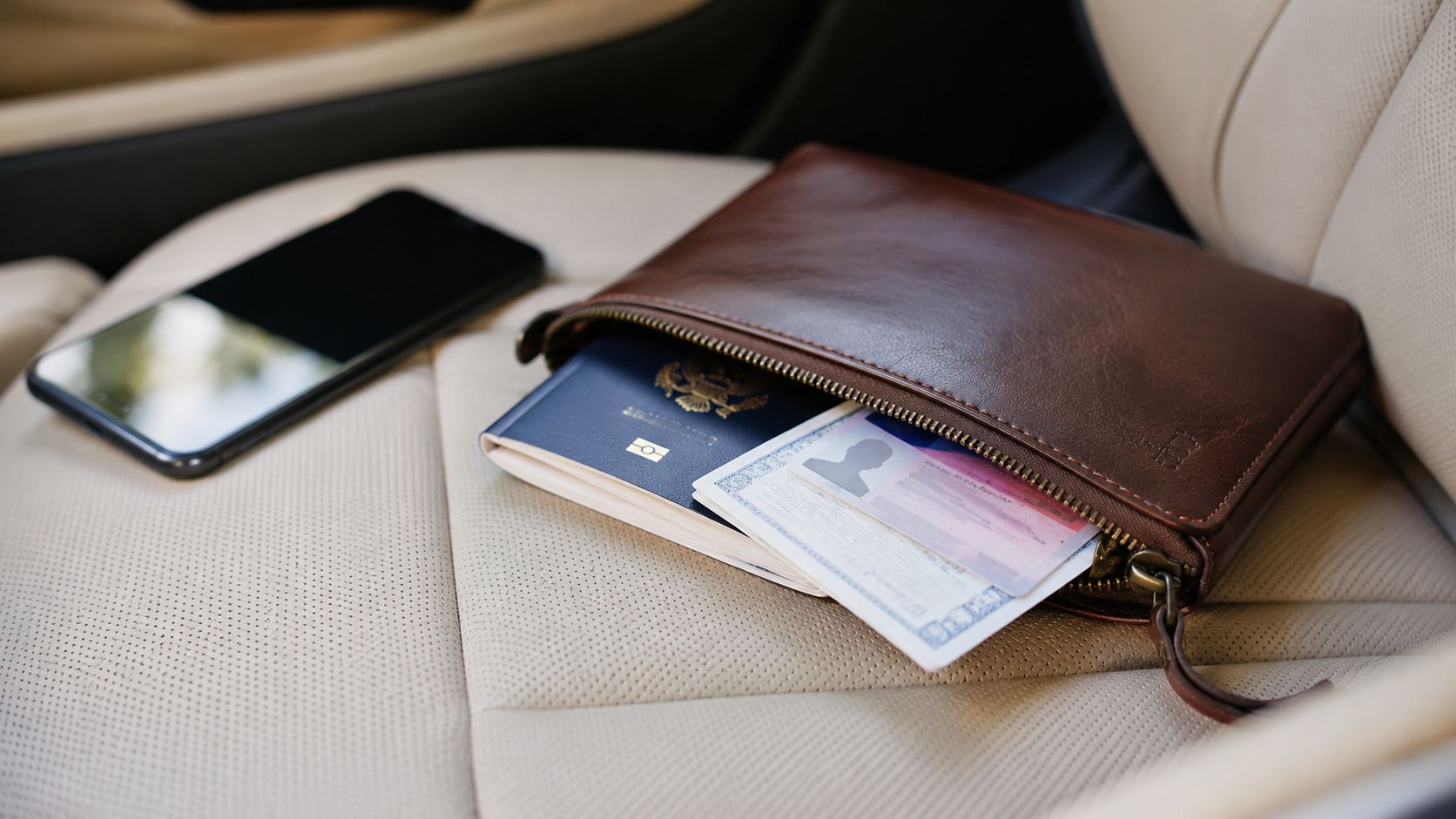 En brun lædermappe indeholder et pas og ID-kort ved siden af en smartphone på et bilsæde.